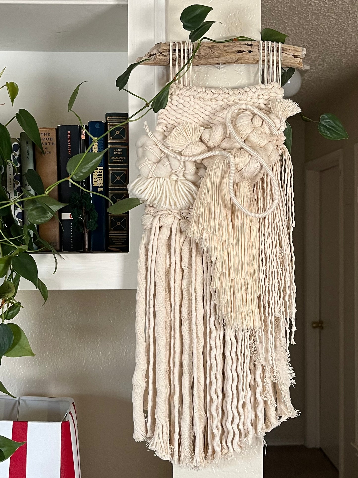 woven wall hanging in a neutral pallete on a white wall with books and plants in the background