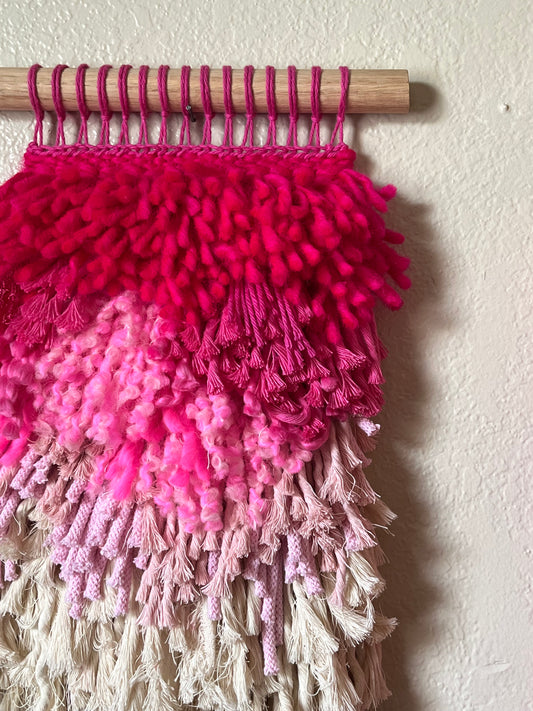 Pink and white textured wall hanging with wooden rod on a white wall
