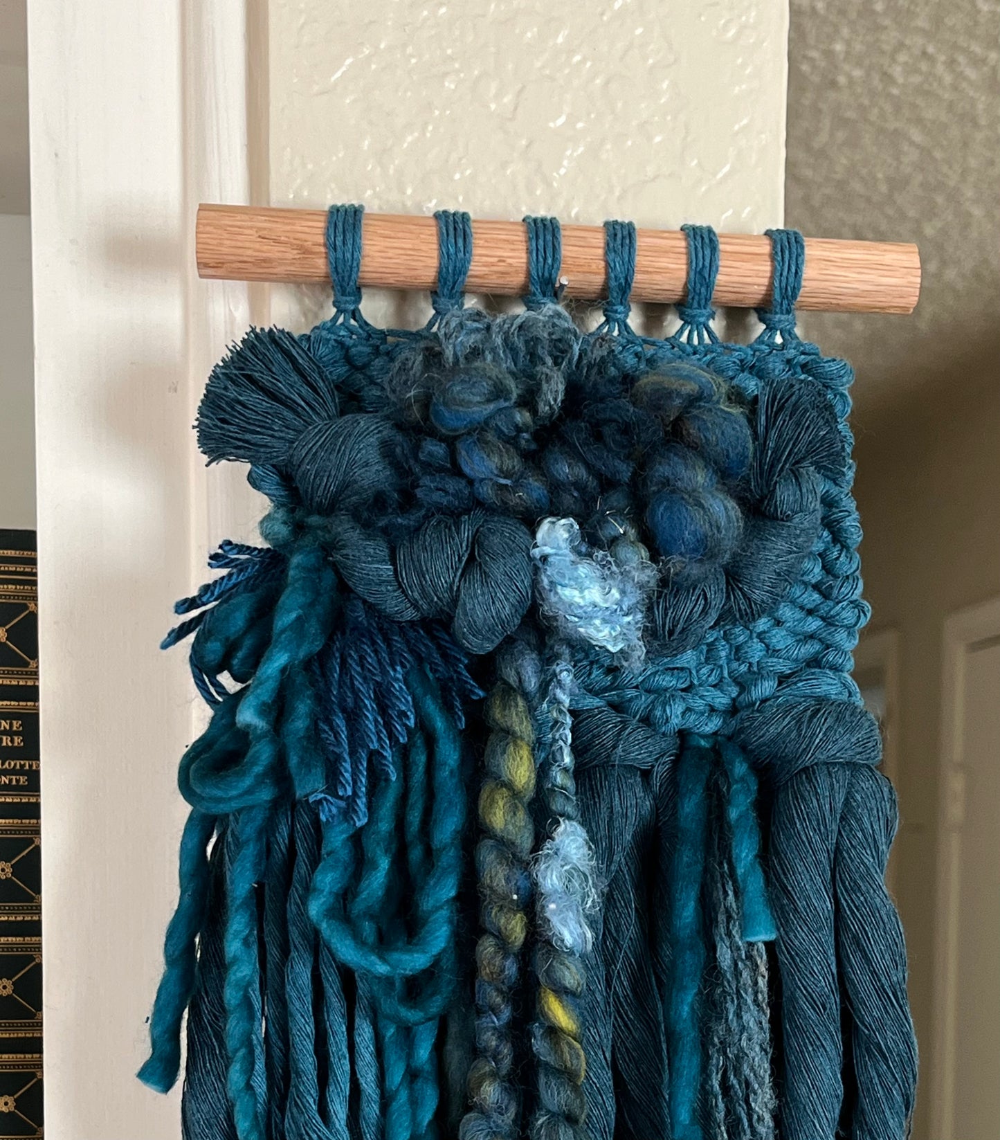 teal monochrome woven wall hanging attached to a wooden rod against a neutral wall.
