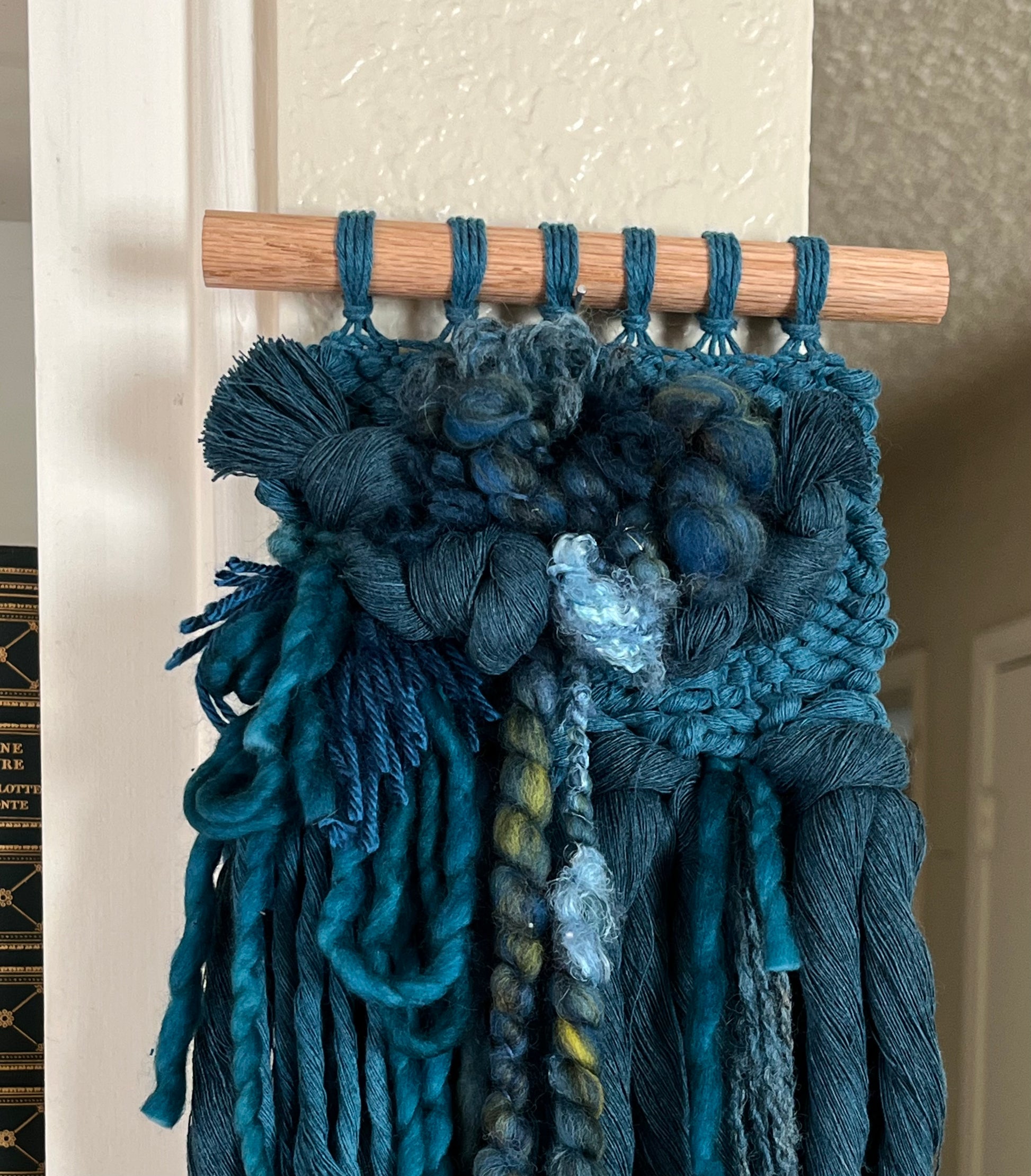 teal monochrome woven wall hanging attached to a wooden rod against a neutral wall.