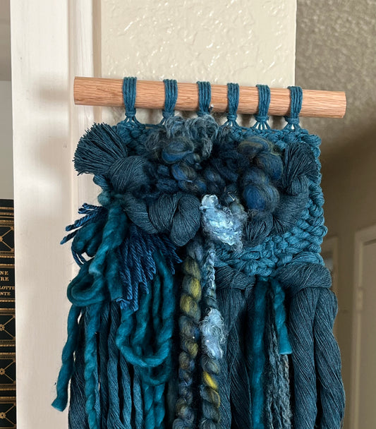 teal monochrome woven wall hanging attached to a wooden rod against a neutral wall.