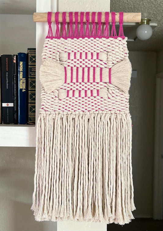 Woven wall hanging with pink and white pattern placed against a neutral background.