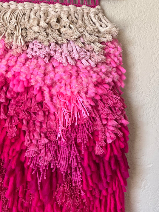 Pink and white textured wall art with fringe hanging on a textured white wall. 